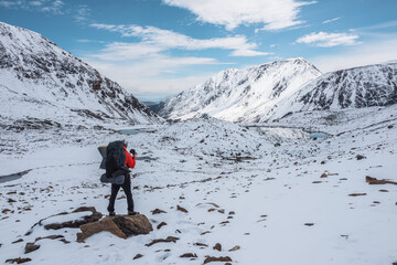 Man in red jacket with big backpack in snowy valley with alpine lakes under clouds in blue sky. Backpacker in snow-covered large mountains among glacial lakes, pure white snow and stones in sunny day. - 790538239