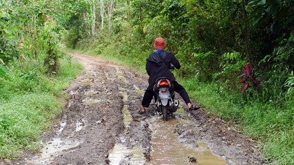 A villager rides a motorbike through a muddy forest road with water and puddles in the village - obrazy, fototapety, plakaty