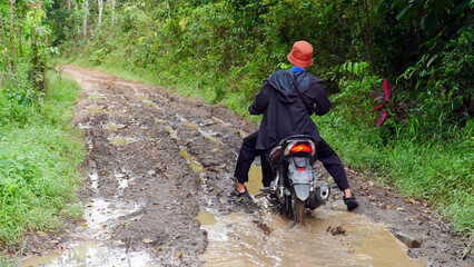 A villager rides a motorbike through a muddy forest road with water and puddles in the village