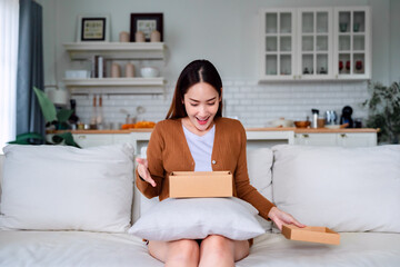 Happy young Asian woman opening delivery package at home. Beautiful female customer open box and sitting on couch in living room