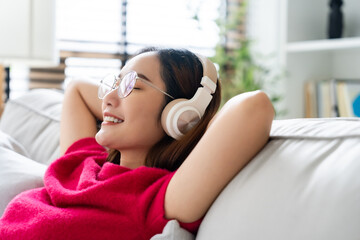 Relaxed young Asian woman enjoying rest on comfortable sofa, calm attractive girl relaxing on couch, breathing fresh air with eyes closed, wearing eyeglasses and headset, meditating at home