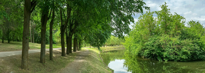 footpath in summer green park along water canal on bright sunny day. panorama.