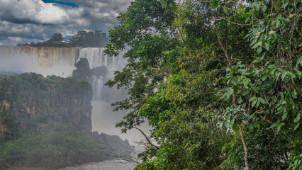 Cascades of waterfalls fall from the cliff, shrouded in spray and fog.  Lush green vegetation in...
