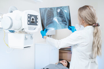 A female doctor looks at an x-ray of the ribs and lungs against the background of x-ray equipment. Diagnosis of Covid, exclusion of pneumonia. Skeleton bones on x-ray