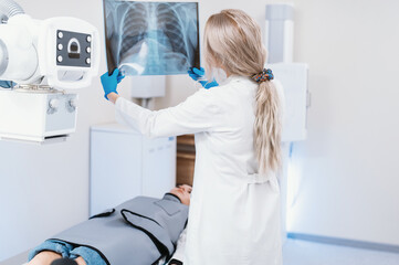 female doctor looks at an x-ray of the ribs and lungs in the radiology room against the background of medical equipment and a lying patient. Fluorography, prevention of pneumonia