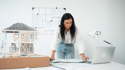Young beautiful caucasian architect puts blueprint on the table and compare with house model while...