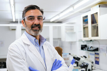 Portrait of a middle aged professional researcher with protective glasses and gloves smiling and looking at camera.