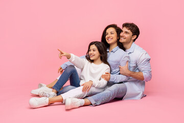 Excited european family of three pointing finger aside, posing sitting on pink studio background,...