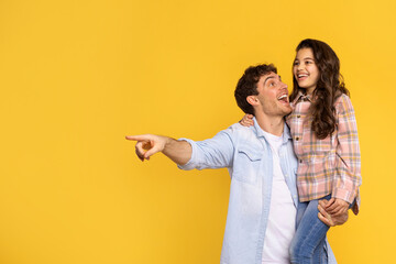 Excited dad holding daughter on hands and pointing aside at free copy space on yellow background,...