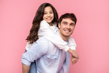 Cheerful father giving his pretty daughter piggyback ride, while they are both looking at camera and smiling over pink background