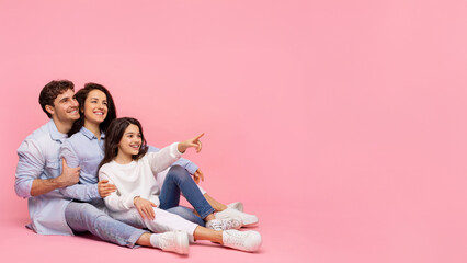 Look there. Excited family of three posing and pointing at free space, sitting on pink background,...