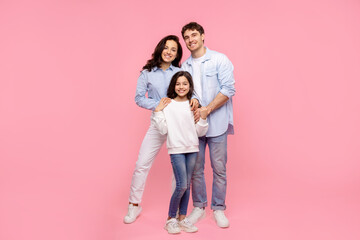 Happy mom dad with child daughter cuddling and smiling at camera, posing isolated on pastel pink background, full length shot