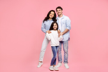 Happy caucasian family of three, parents and their pretty daughter, posing on plain pastel pink...