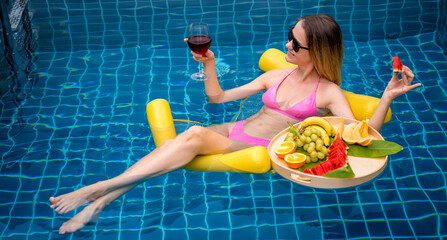 Beautiful woman lying on floating hammock in the swimming pool with wine and tray of fruits