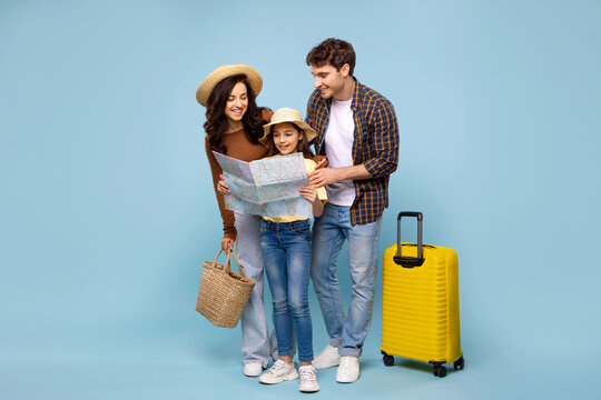 Happy family with daughter looking at map, planning travel, standing with suitcase on blue studio background, ready for summer vacation