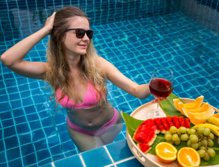 Beautiful woman in the swimming pool with glass of wine and floating tray of fruits - 790602893