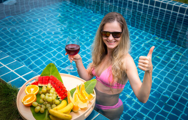 Beautiful woman in the swimming pool with glass of wine and floating tray of fruits - 790603297