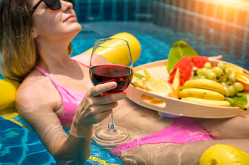 Beautiful woman lying on floating hammock in the swimming pool with wine and tray of fruits - 790604601