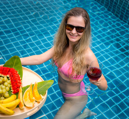 Beautiful woman in the swimming pool with glass of wine and floating tray of fruits - 790606685