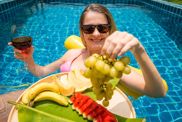 Beautiful woman lying on floating hammock in the swimming pool with wine and tray of fruits - 790611683