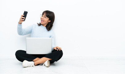Young mixed race woman sitting on the floor with laptop isolated on white background making a selfie - Powered by Adobe