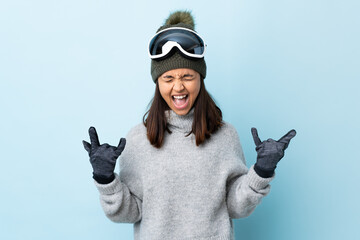 Mixed race skier girl with snowboarding glasses over isolated blue background making horn gesture.