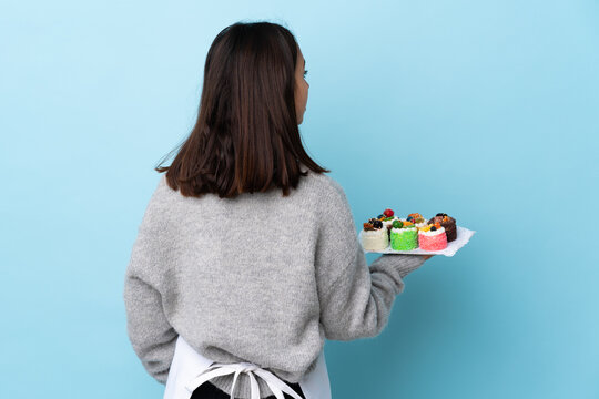 Pastry chef holding a big cake over isolated blue background in back position and looking back.