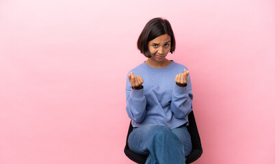 Young mixed race woman sitting on a chair isolated on pink background making money gesture but is...