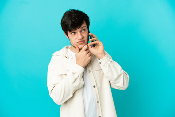 Young Russian man using mobile phone isolated on blue background having doubts