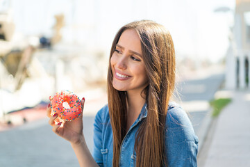 Young woman holding a donut at outdoors looking up while smiling