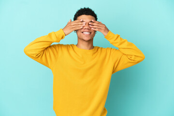 Young African American man isolated on blue background covering eyes by hands and smiling