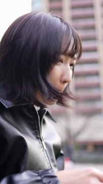Vertical slow-motion video of a Japanese woman in her 20s sitting and putting on makeup in a park around Gotanda Station, Shinagawa-ku, Tokyo in winter 冬の東京都品川区五反田の駅周辺の公園で座って化粧をする２０代の日本人女性の縦長スローモーション動