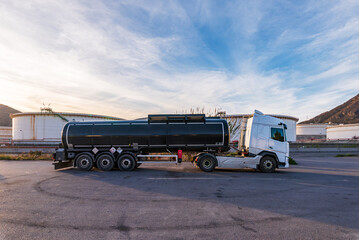 Tanker truck for transporting dangerous goods next to large oil storage tanks, side view.
