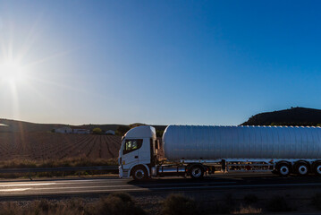 Gas tanker truck driving on a conventional road with a sun at dawn, side view.