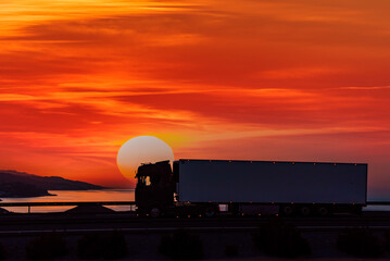 Refrigerator truck driving by the sea with a huge sun at dawn and a completely red sky, side view. Hard and painful profession rewarded with wonderful sunrises.
