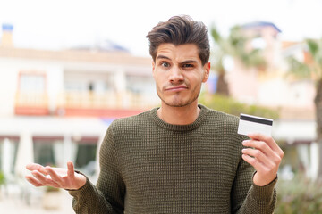 Young caucasian man holding a credit card at outdoors making doubts gesture while lifting the...