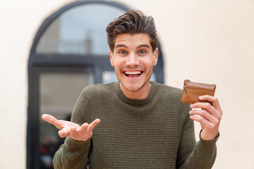 Young caucasian man holding a wallet at outdoors with shocked facial expression