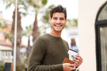 Young caucasian man at outdoors holding wallet and credit card with happy expression