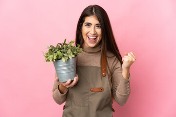 Young gardener girl holding a plant isolated on pink background celebrating a victory in winner...