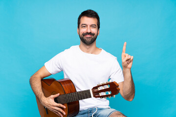 Young man with guitar over isolated blue background showing and lifting a finger in sign of the best