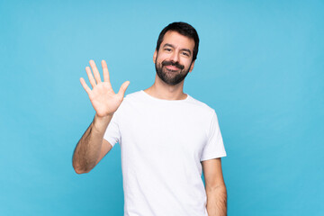 Young man with beard  over isolated blue background saluting with hand with happy expression