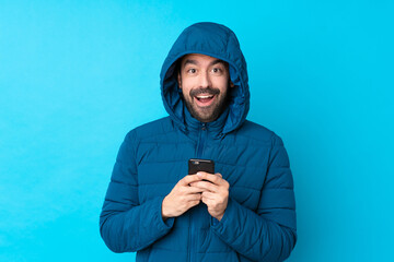 Man wearing winter jacket and holding a takeaway coffee over isolated blue background surprised and sending a message