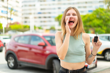 Young blonde woman holding car keys at outdoors shouting with mouth wide open
