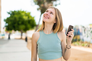 Young blonde woman using mobile phone at outdoors looking up while smiling