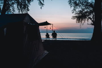 man and a teddy bear sit on a chair having a rest in front of tent at campfire under morning sky,...
