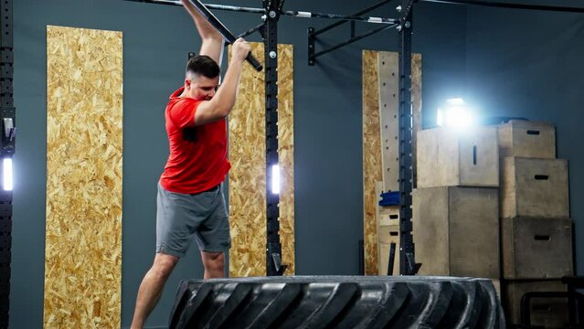 Athlete hits big tire with hammer. Concept of strength and determination. Sportsman training in modern gym. Man in red shirt standing in front of tire, preparing to hit it with hammer. 4K, UHD