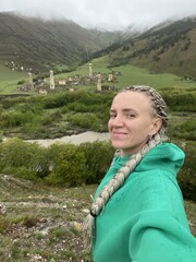 Naklejka premium A girl on the background of the medieval Targim tower complex in the Caucasus mountains, surrounded by greenery. The Caucasus Mountains on a cloudy day in Ingushetia. Russia