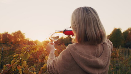 A woman pours red wine into a glass. It stands near the vineyard at sunset. Tasting and wine tour concept