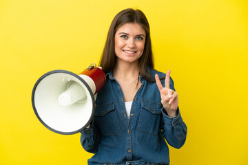 Young caucasian woman isolated on yellow background holding a megaphone and smiling and showing...