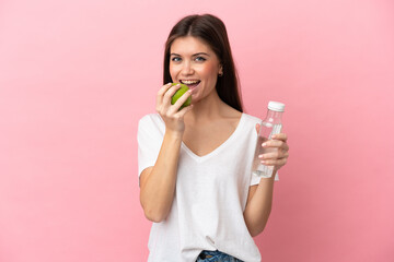 Young caucasian woman isolated on pink background with a bottle of water and eating an apple